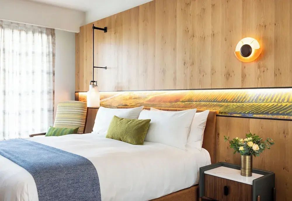 Doppelbett im Hotelzimmer mit Holzrückwand, beigefarbenem Sessel und kleiner Pflanze.