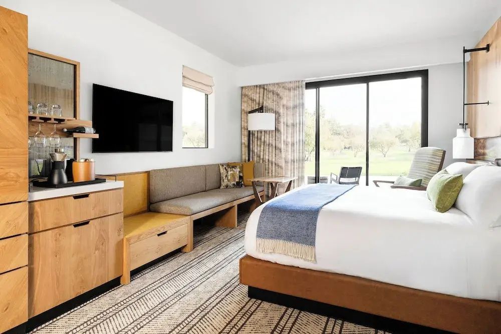Helles Hotelzimmer mit Doppelbett, hölzernen Einbaumöbeln, Wandfernseher und Blick ins Grüne.