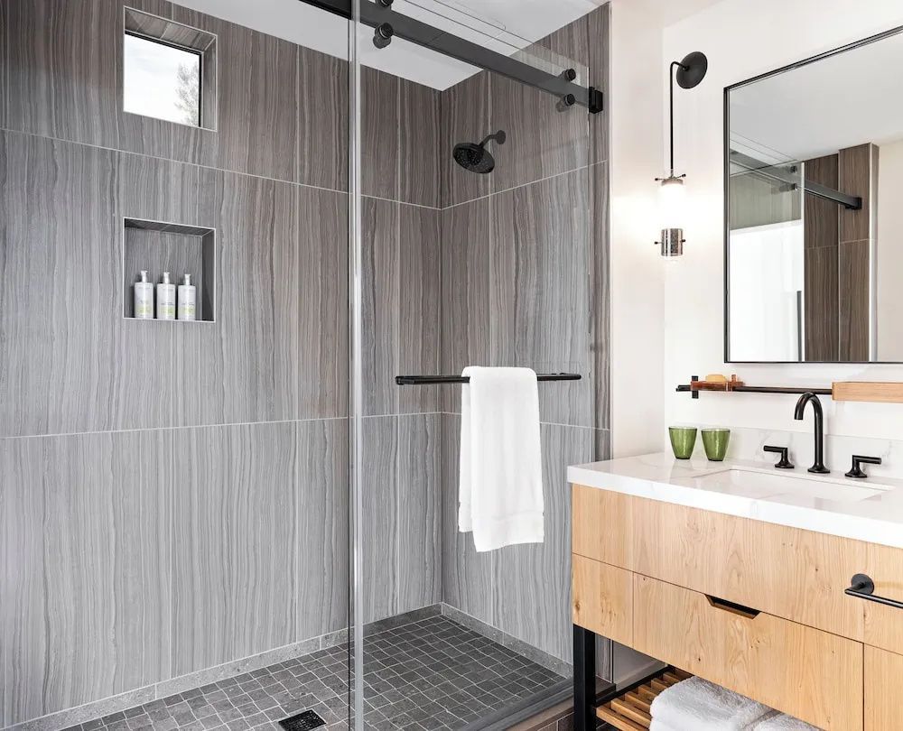 Walk-in-Dusche mit grauen Wandfliesen und Holzwaschtisch im hellen Badezimmer.