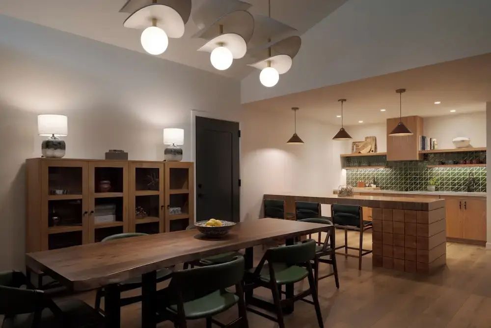 Langer Holzesstisch mit grünen Stühlen, Vitrinenschrank und offener Küchenbereich mit Bar.
