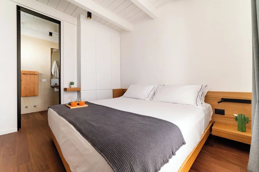 Doppelbett mit weißer Bettwäsche, grauer Tagesdecke und Holzrahmen in einem hellen Zimmer.