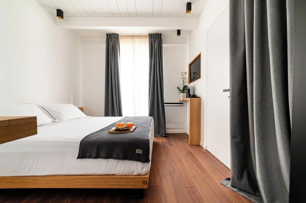 Schlafzimmer mit Doppelbett, Parkettboden, bodentiefem Fenster und grauen Vorhängen.