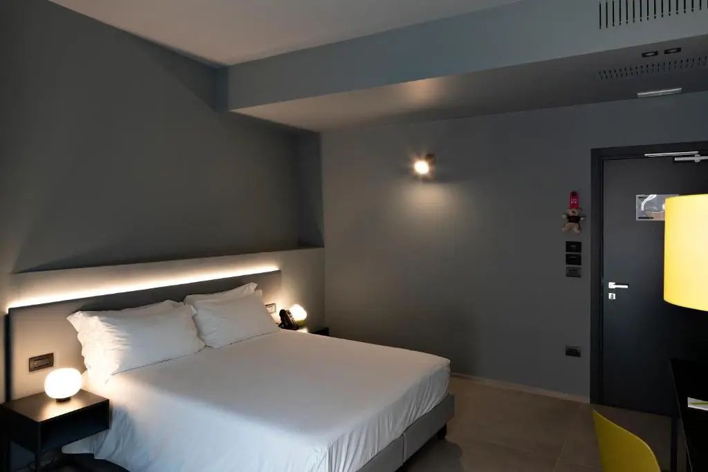 Doppelbett mit weißer Bettwäsche und beleuchtetem Kopfteil vor grauen Wänden.