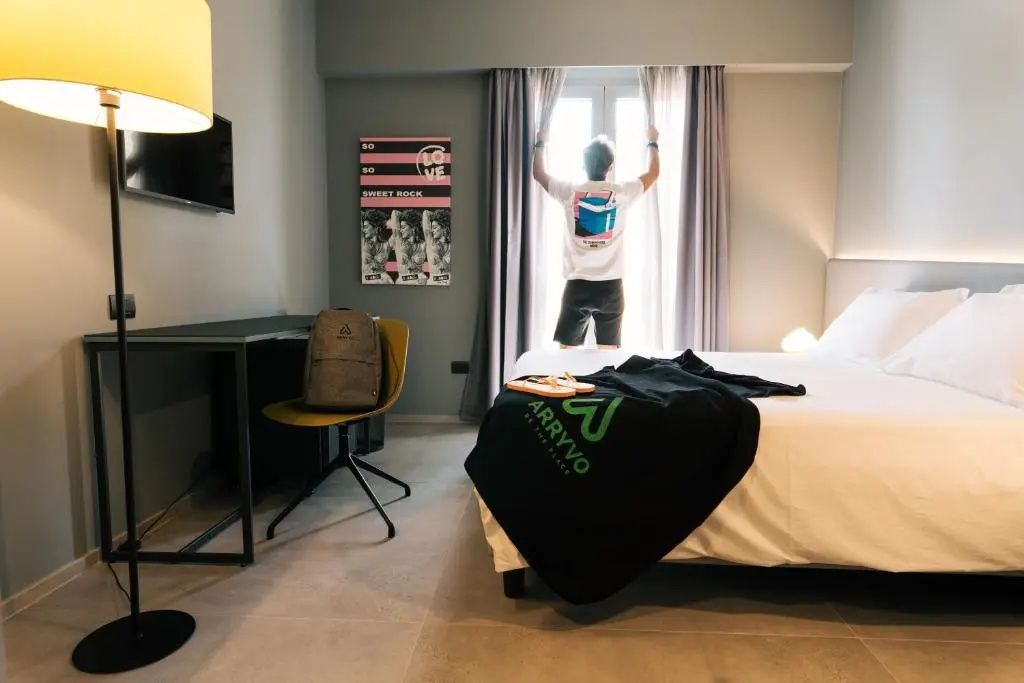 Ein Hotelzimmer mit Bett, Schreibtisch und einer Person am hellen Fenster.