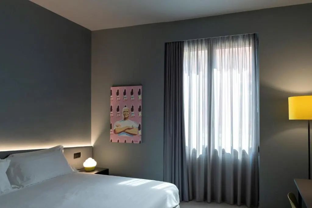 Blick auf das Bett im Hotelzimmer mit grauer Wand, Kunstwerk und Vorhang.