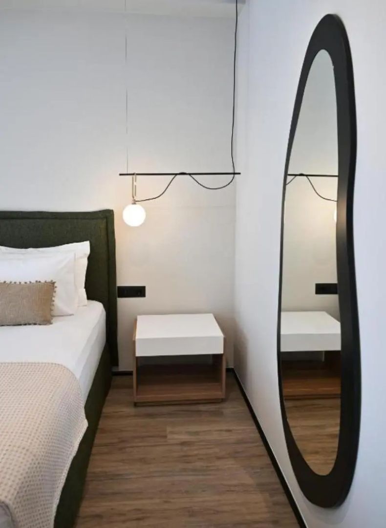 Bett mit grünem Kopfteil, weißem Nachttisch, Hängeleuchte und einem geschwungenen Wandspiegel.
