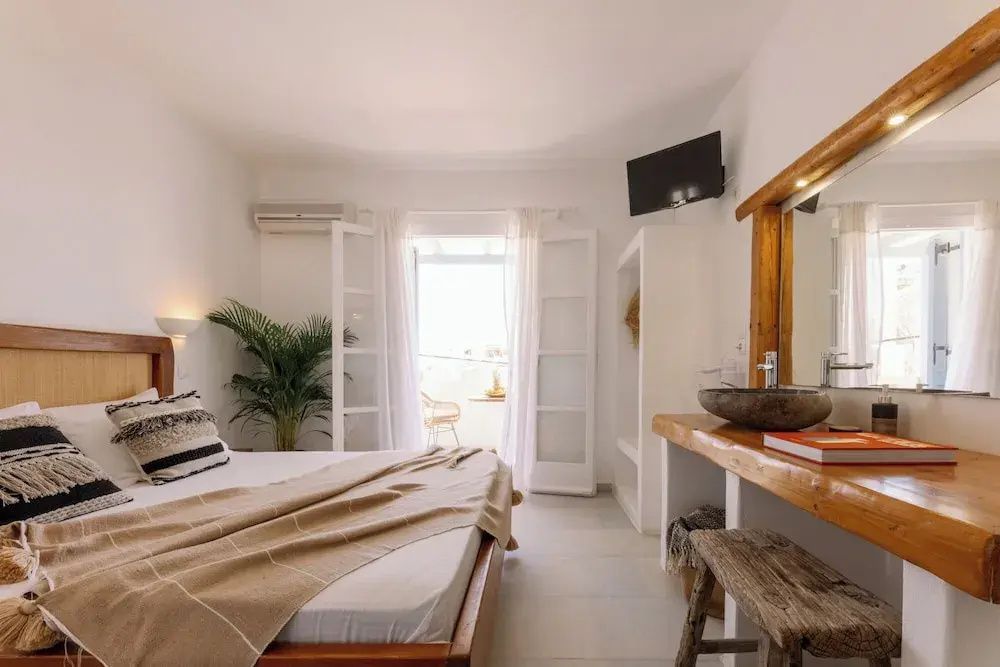 Helles Zimmer mit Holzdoppelbett, Steinwaschbecken am Tisch und offener Balkontür.