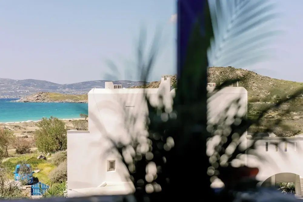 Blick vorbei an weißen Gebäudefronten auf das blaue Meer und die hügelige Küstenlandschaft.