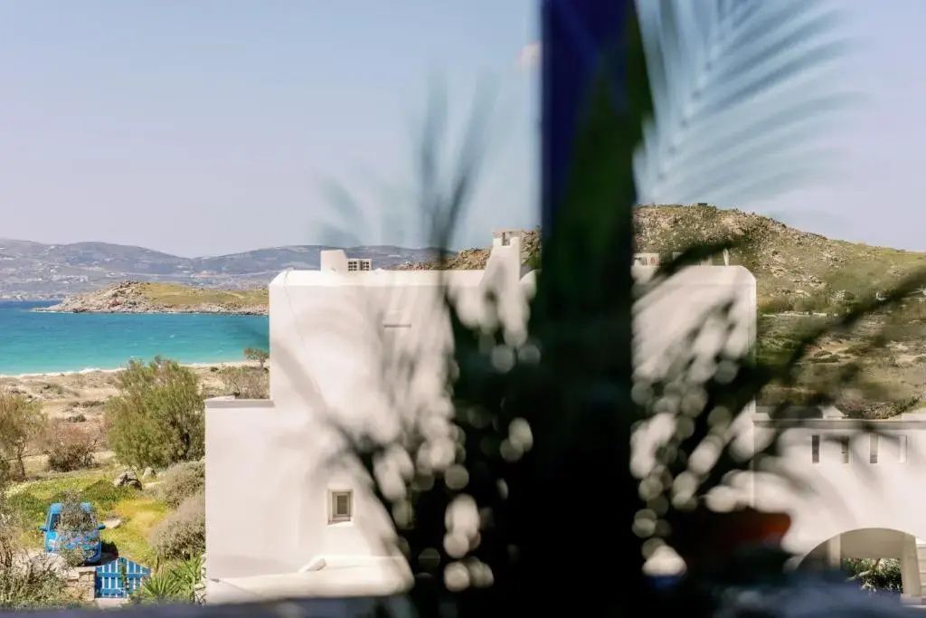 Blick an einem weißen Gebäude vorbei auf eine blaue Meeresbucht und hügelige Küstenlandschaft.
