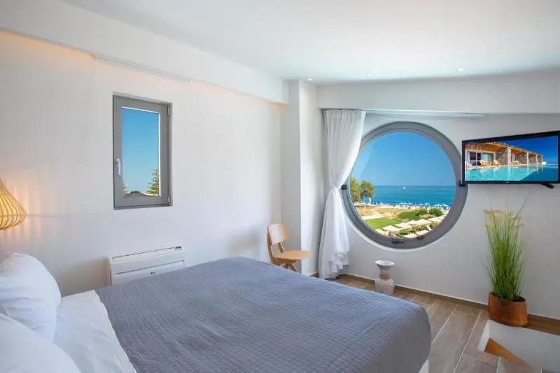 Helles Schlafzimmer mit Doppelbett und einem großen runden Fenster mit Blick auf das Meer.