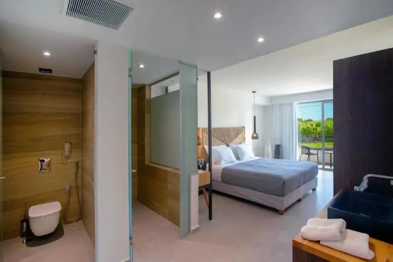 Doppelbett in hell eingerichtetem Hotelzimmer mit angrenzendem Duschbereich aus Glas und Holz.