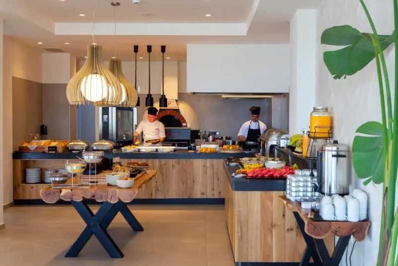 Restaurantbereich mit Buffet, offener Küche, Holzmöbeln und zwei Köchen bei der Arbeit.