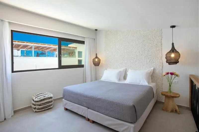 Doppelbett mit grauer Tagesdecke vor einer strukturierten Wand und Fenster.