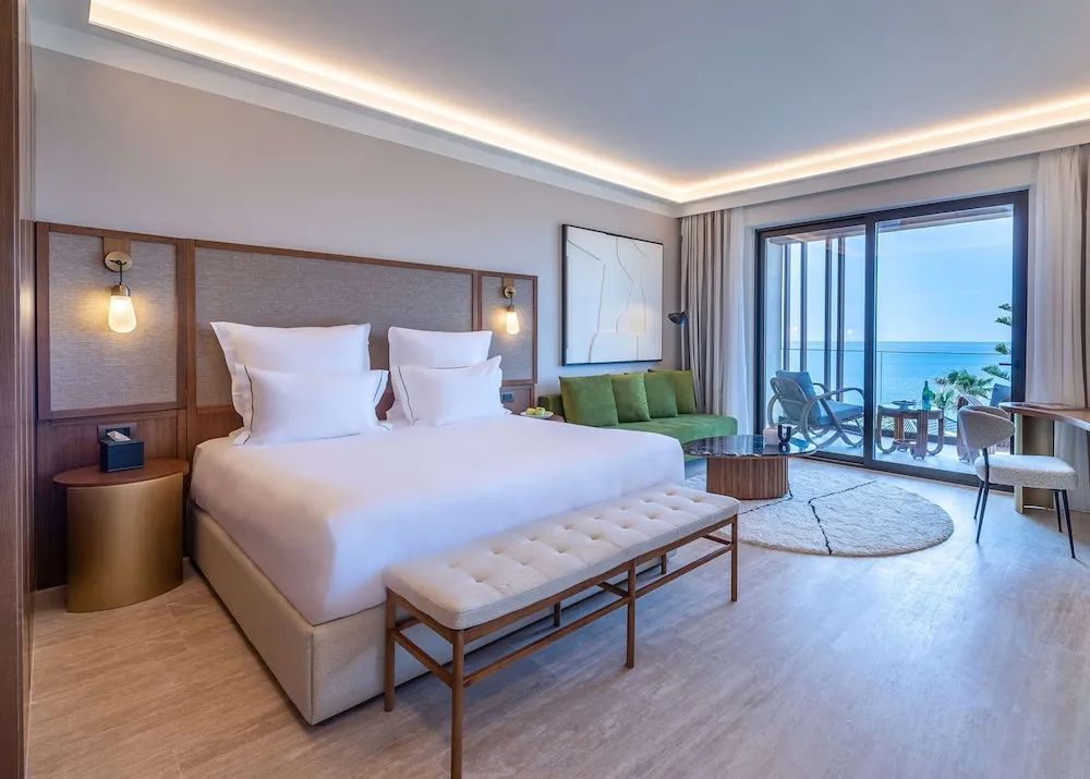 Modernes Hotelzimmer mit grossem Doppelbett, grünem Sofa und Balkon mit Meerblick.