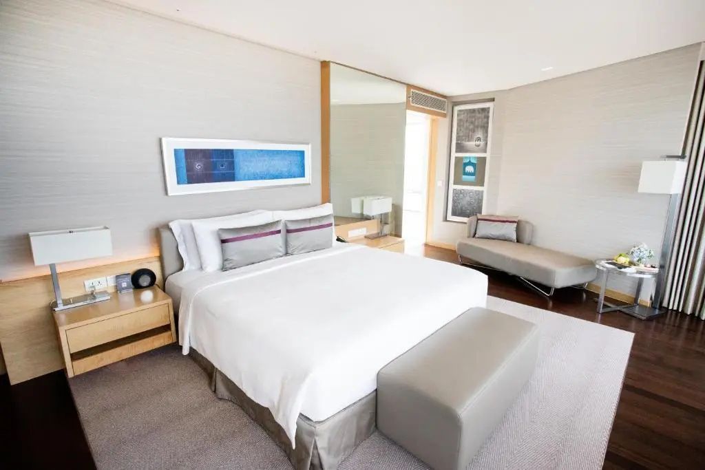 Doppelbett mit weißer Bettwäsche, grauer Sitzbank und Liege in einem hellen Hotelzimmer.