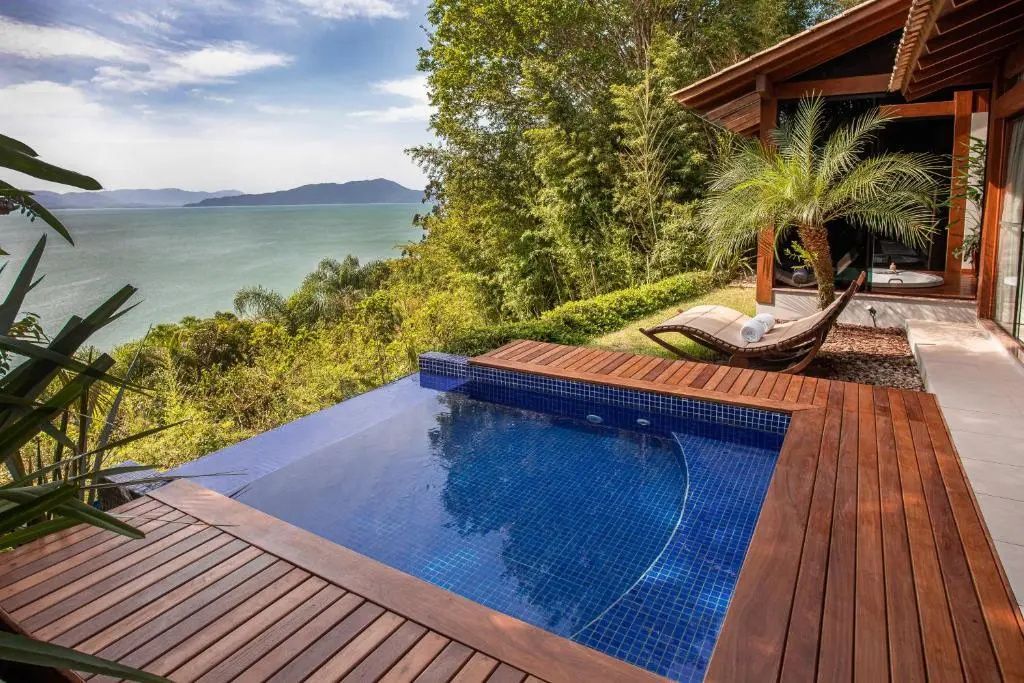 Holzdeck mit kleinem Pool und Liegestuhl vor grüner Vegetation und Blick aufs Meer.