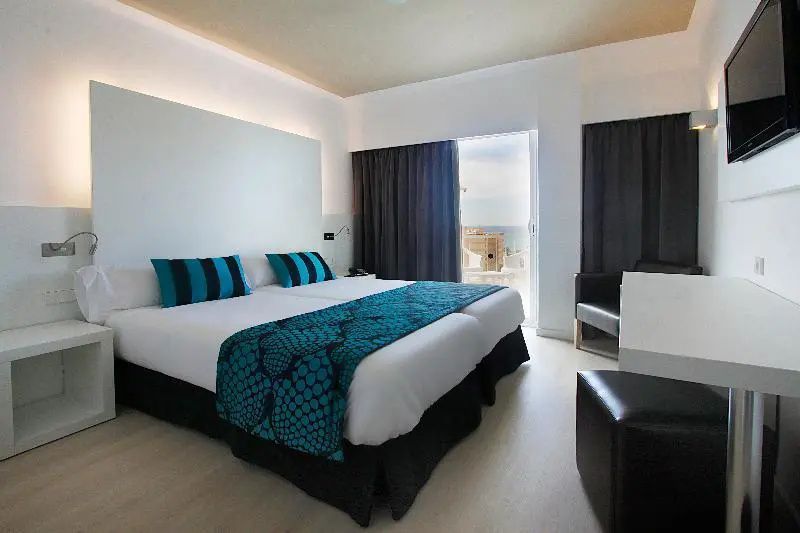 Doppelbett mit türkisblauer Tagesdecke und Kissen vor geöffneter Balkontür mit Meerblick
