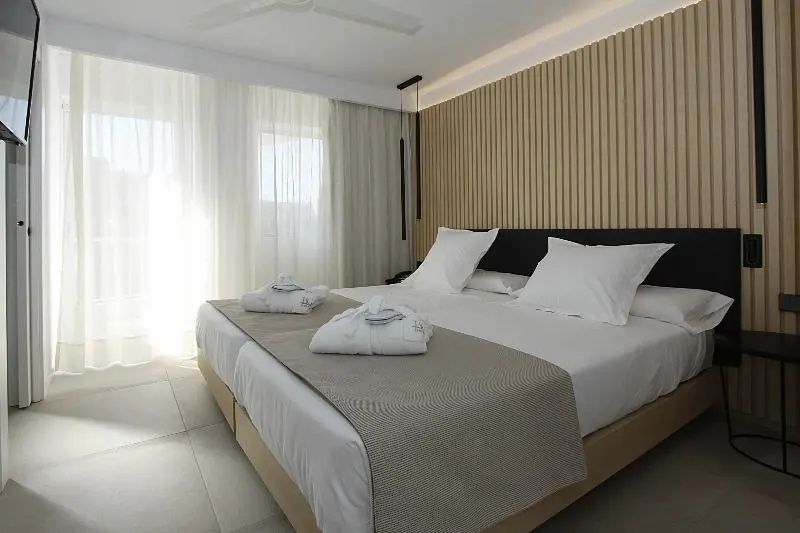 Doppelbett mit weißen Bademänteln vor einer Wand aus Holzlamellen im Hotelzimmer.