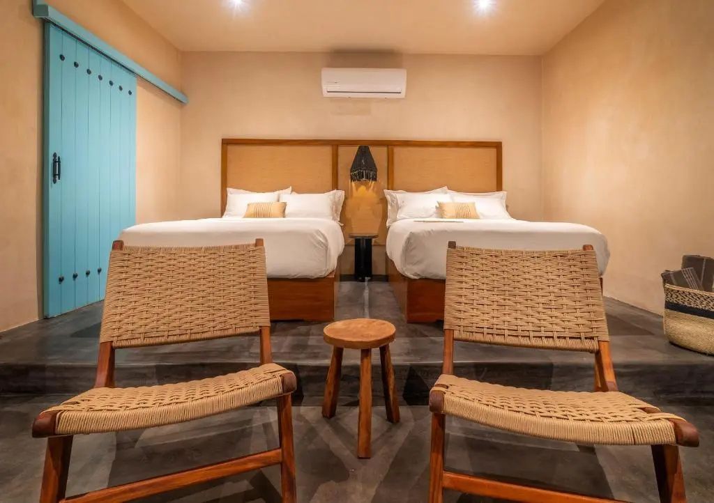 Zwei Doppelbetten und zwei Flechtsessel aus Holz in einem hell eingerichteten Hotelzimmer.