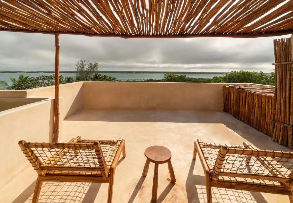 Dachterrasse mit Überdachung aus Holzästen, zwei Holzstühlen und Blick auf die Lagune.