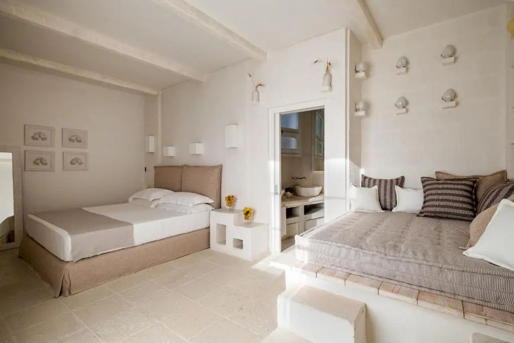 Hell eingerichtetes Hotelzimmer mit Doppelbett, Tagesbett und Blick ins angrenzende Badezimmer.