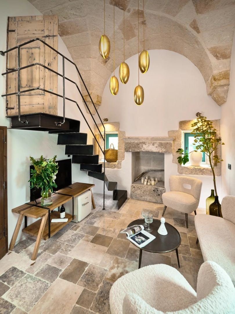 Wohnbereich einer Suite mit Kamin aus Stein, Treppe, hellen Sesseln und goldenen Hängeleuchten.
