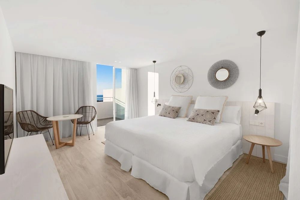 Schlafzimmer mit weißem Doppelbett, Sitzecke und Balkon mit Ausblick auf das Meer.