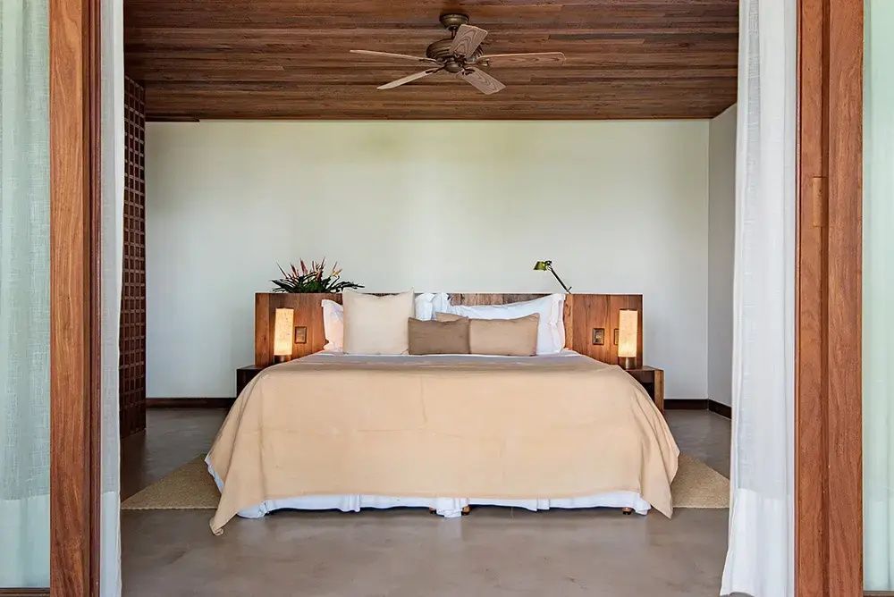 Doppelbett mit beiger Tagesdecke, Holzkopfteil und Deckenventilator unter einer Holzdecke.