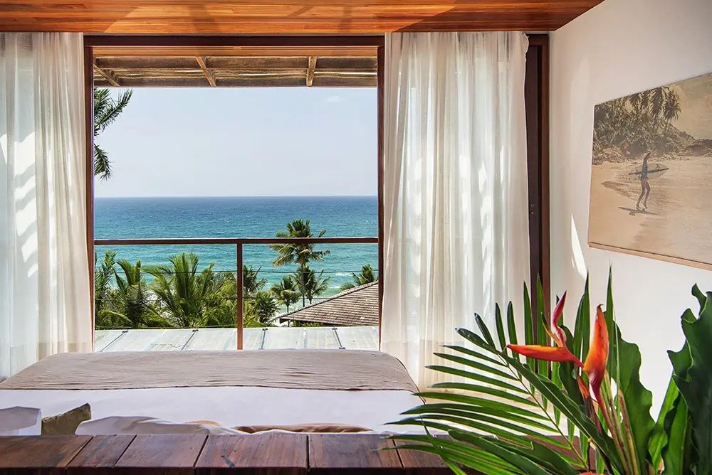 Blick vom Bett durch die geöffnete Balkontür auf Palmen und das blaue Meer.