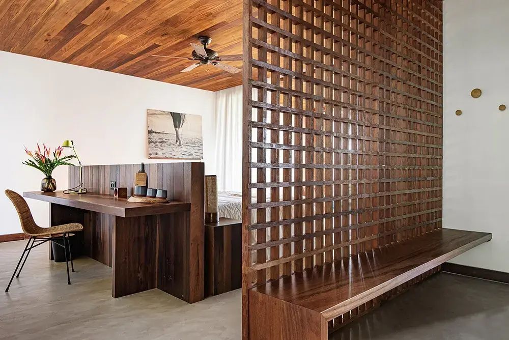 Holzgitter als Raumteiler neben einer Holzbank in einem hell eingerichteten Hotelzimmer.