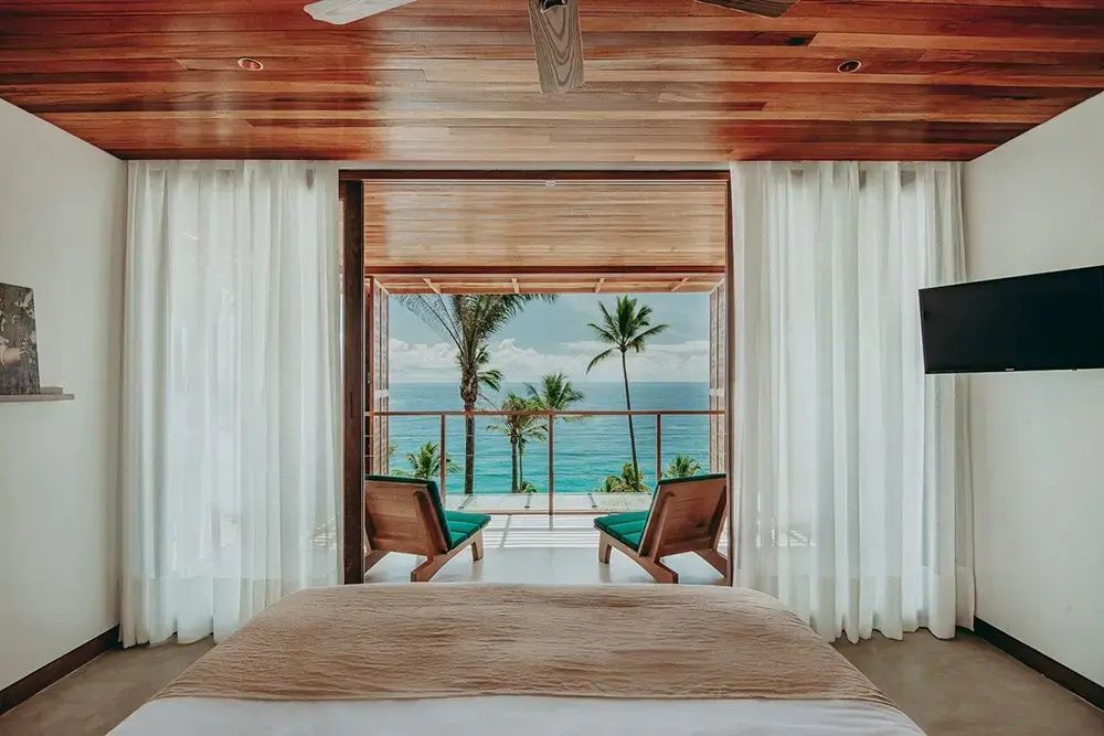 Blick vom Bett durch eine offene Glasfront auf den Balkon, Palmen und das Meer.