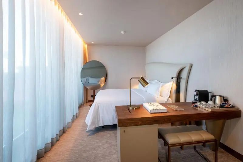 Helles Hotelzimmer mit Doppelbett, hölzernem Schreibtisch und großem runden Spiegel.