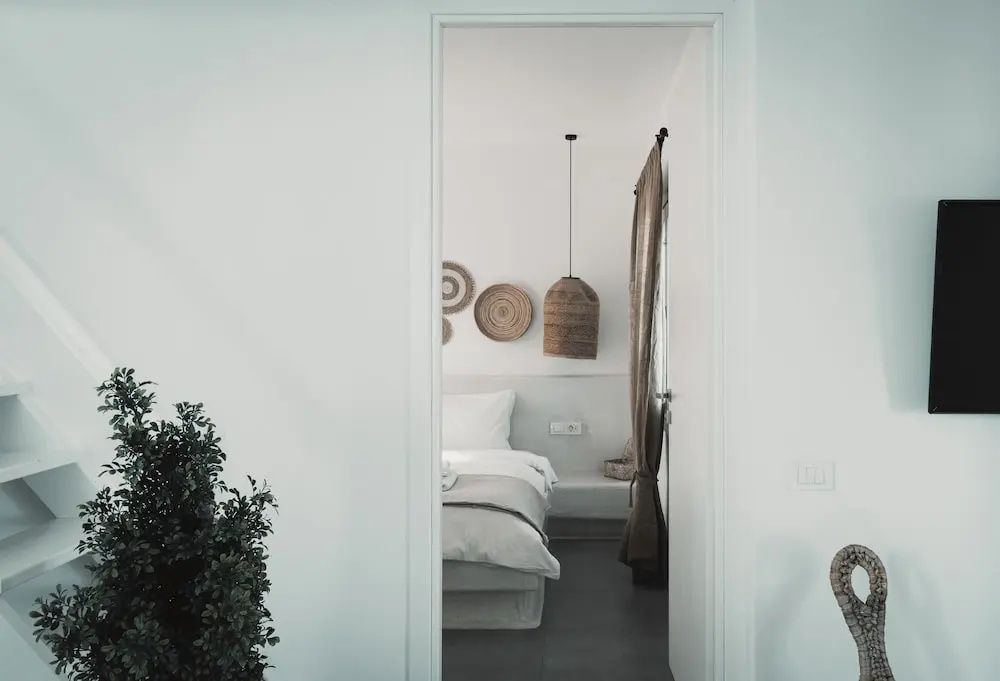 Blick durch eine weiße Türöffnung auf ein hell eingerichtetes Schlafzimmer mit Bett und Rattanlampe.