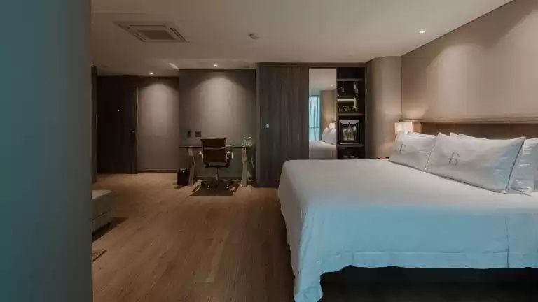 Hotelzimmer mit weißem Doppelbett, Parkettboden und Schreibtisch im Hintergrund.