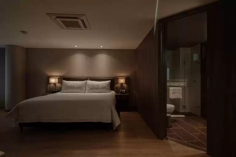 Gepflegtes Doppelbett mit weißer Bettwäsche, beleuchteten Nachttischen und Blick ins Badezimmer.