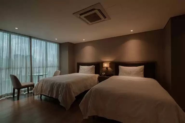 Hotelzimmer mit zwei Einzelbetten, Nachttisch und großem Fenster mit hellen Vorhängen.