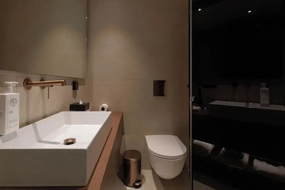 Modernes Badezimmer mit weißem Waschtisch auf Holzkonsole, WC und bronzenem Abfalleimer.