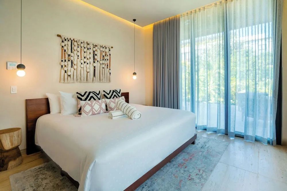 Doppelbett mit gemusterten Kissen, Makramee Wandbehang und Fenster zur Terrasse.