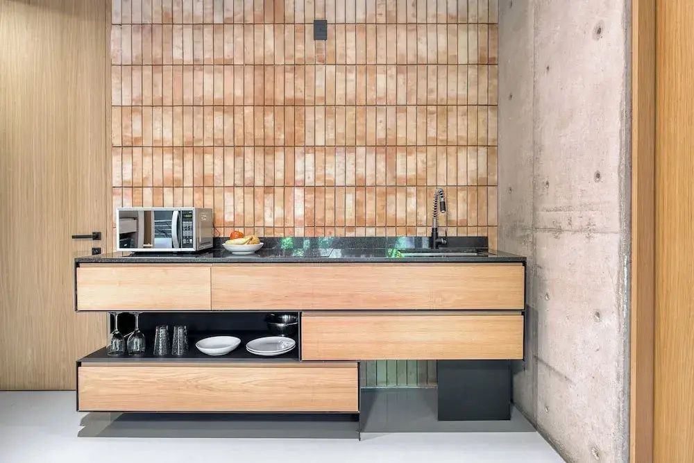 Modernes Kitchenette-Element mit Holzfronten, dunkler Arbeitsplatte und einer Wand aus schmalen Ziegeln.