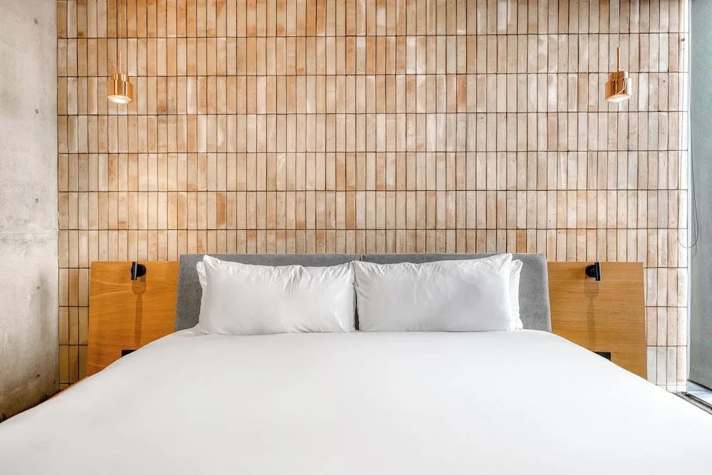 Doppelbett mit weißer Bettwäsche vor einer dekorativen Wand aus schmalen Fliesen.