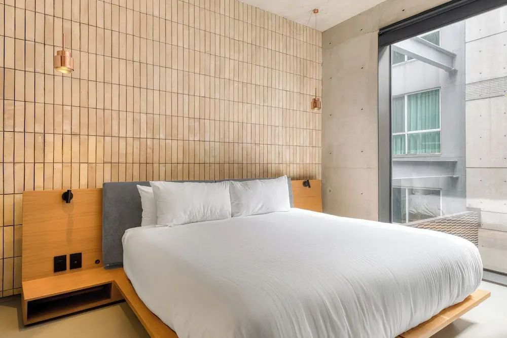 Doppelbett mit weißer Bettwäsche vor einer hellen Fliesenwand neben einem großen Fenster.
