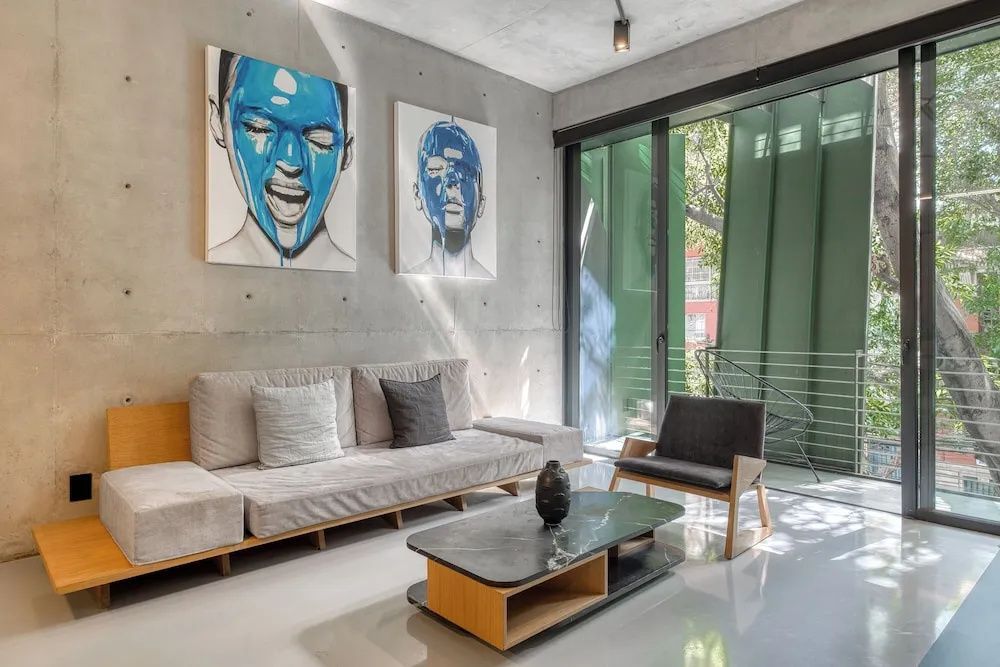 Helles Sofa mit Kissen neben einem Couchtisch und Sessel vor Sichtbetonwand mit blauen Kunstwerken.