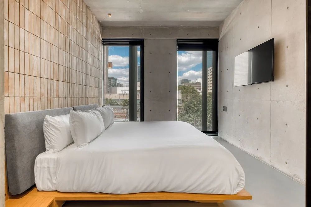 Helles Schlafzimmer mit großem Holzdoppelbett, Fliesenwand und zwei hohen Fenstern.