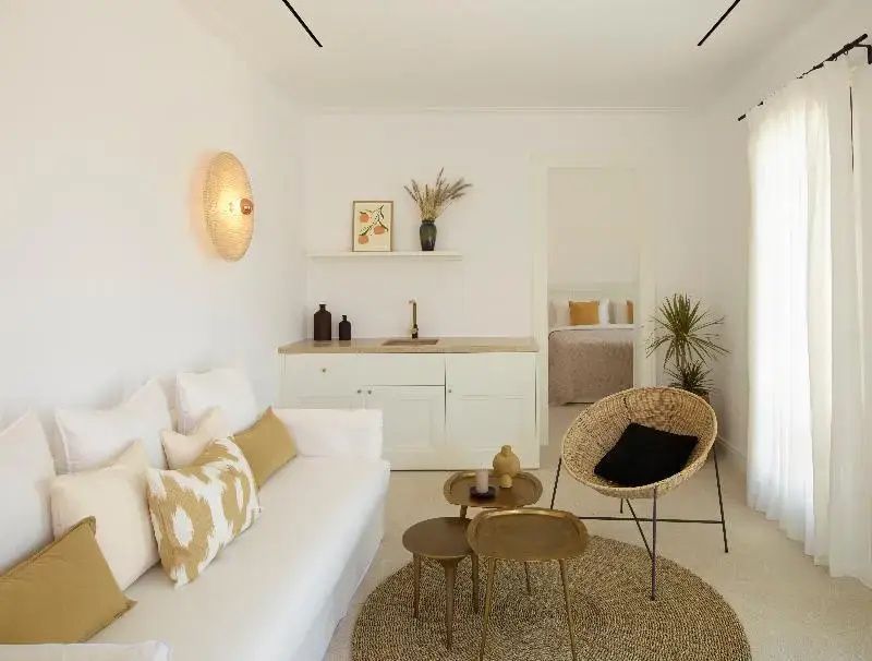 Heller Wohnbereich mit weißem Sofa, Flechtstuhl und einer kleinen Küchenzeile im Hintergrund.