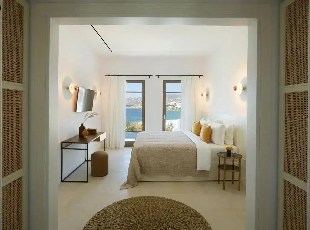 Hell eingerichtetes Schlafzimmer mit Doppelbett und Blick auf das Meer durch zwei Fenster.