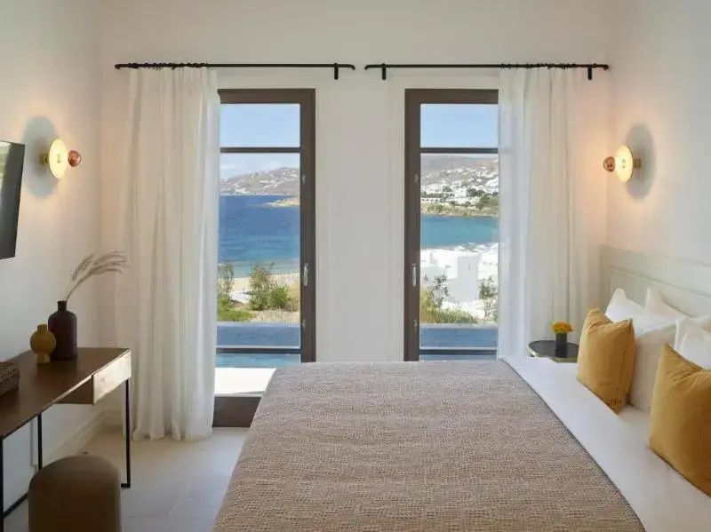 Blick durch zwei hohe Fenster im Schlafzimmer auf das blaue Meer und die Küste.