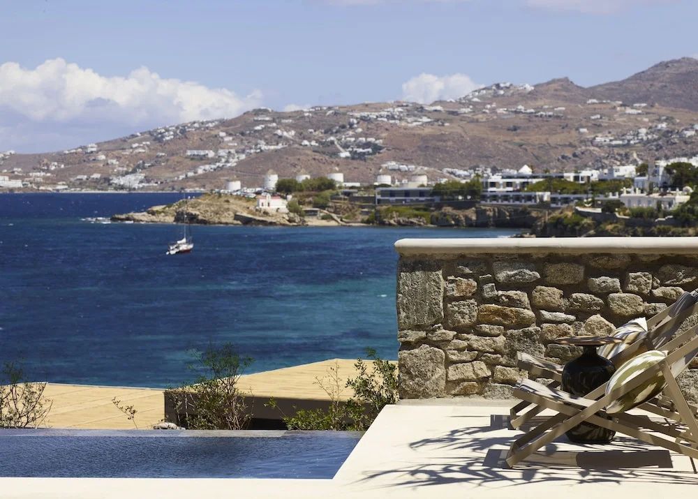 Terrasse mit Steinmauer und Blick über das blaue Meer auf die Küste von Mykonos.