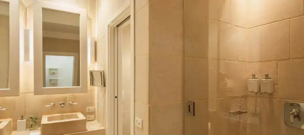 Badezimmer mit hellem Naturstein, Glastür zur Dusche und einem Kosmetikspiegel.