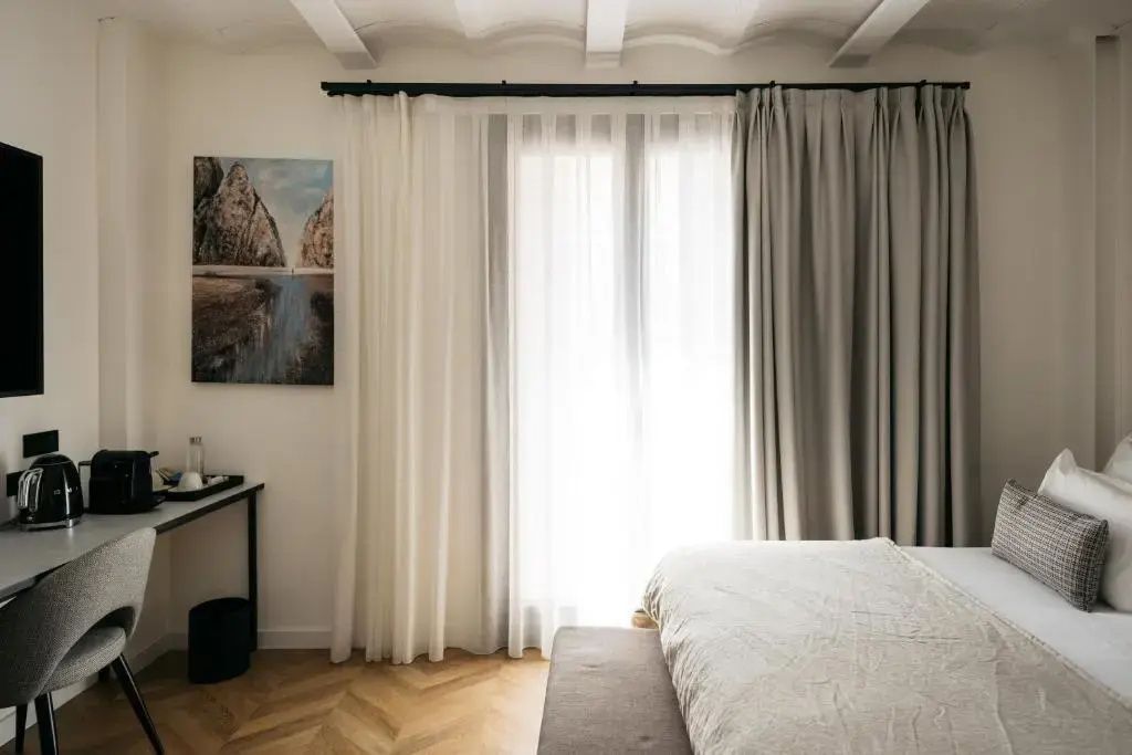 Helles Hotelzimmer mit bodentiefen Vorhängen, Parkettboden und einem Bett.