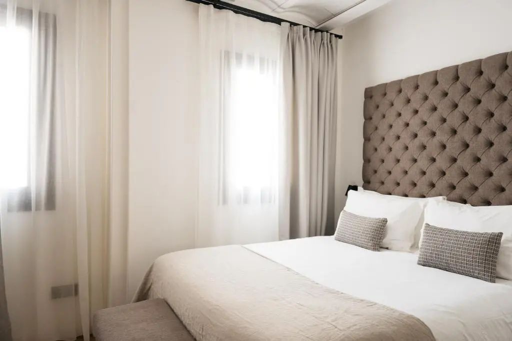 Doppelbett mit gepolstertem Kopfteil und hellem Vorhang am Fenster im Hotelzimmer.